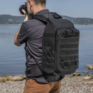 Tenba-axis-V2-20L-7 Tenba Axis V2 20L -reppu -kamerareppu mukanaan kantava henkilö seisoo kivikkoisella rantaviivalla ja ottaa kuvia tyynestä vedestä ja kaukaisista kukkuloista.