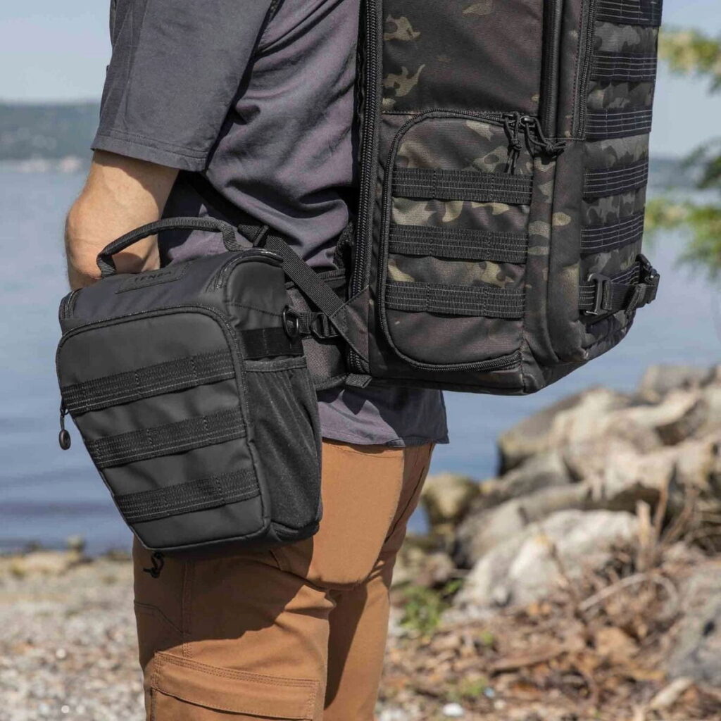 Tenba-axis V2-4L-7 Out on the water, a person wearing brown trousers and a grey shirt carries a Tenba Axis V2 4L Top Loader and a removable black bag on his hip, with rocks and greenery in the background.