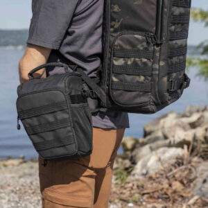 Tenba-axis V2-4L-7 Out on the water, a person wearing brown trousers and a grey shirt carries a Tenba Axis V2 4L Top Loader and a removable black bag on his hip, with rocks and greenery in the background.