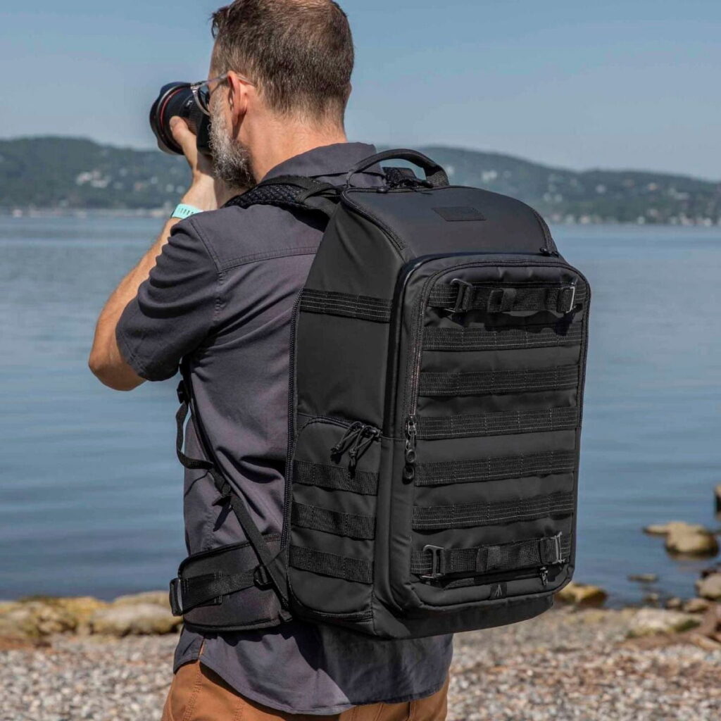 Tenta-axis V2-9 A short, bearded man in a dark shirt and carrying a Tenba Axis V2 24L Backpack is standing on a rocky beach taking photos. Water and distant hills can be seen in the background.