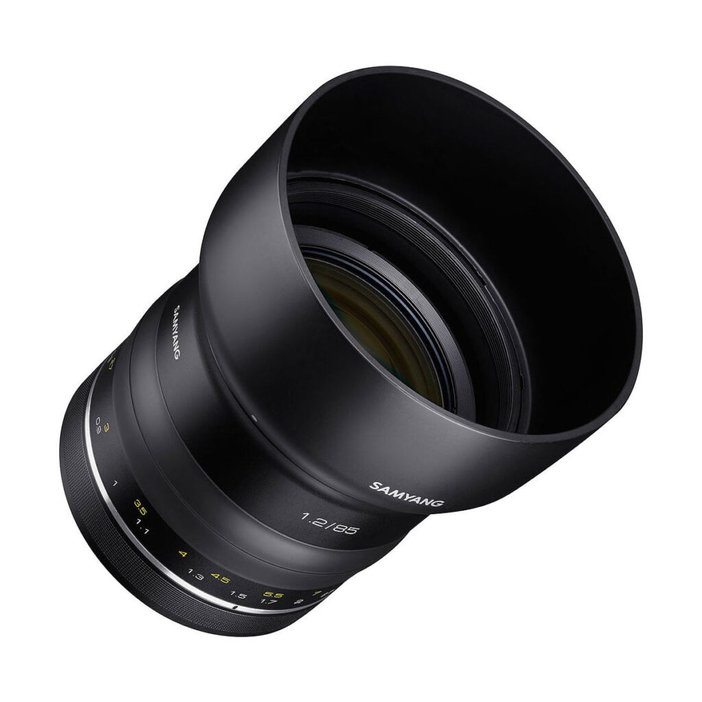 Samyang_XP_85mm_f_1_2_003 Samyang XP 85mm f/1.2 for Canon EF on musta kameraobjektiivi, jossa on keltaiset ja valkoiset merkinnät, leveä huppu ja aukkomerkinnät, kuvattuna kulmassa valkoisella pohjalla.