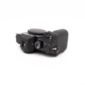 Fujifilm X-S10-3 Fujifilm X-S10 (SC 1189) - Käytetty digitaalikameran runko näkyy ylhäältä valkoisella pohjalla, ja siinä korostuvat toimintatilan valitsin, virtakytkin, painikkeet ja objektiivin kiinnityksen peittävä suojakansi.