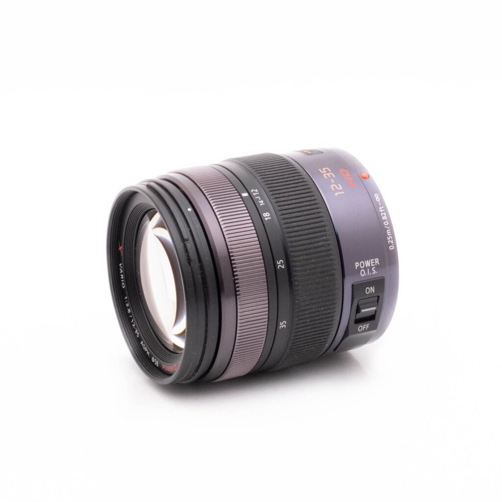 Lumix Vario 12-35-2 Musta Panasonic Lumix Vario 12-35mm f/2.8 Power O.I.S - Käytetty -objektiivi näkyy valkoisella pohjalla vasemmalle suunnattuna, ja siinä näkyvät tarkennus- ja zoomausrenkaat sekä Power O.I.S. -kytkin.