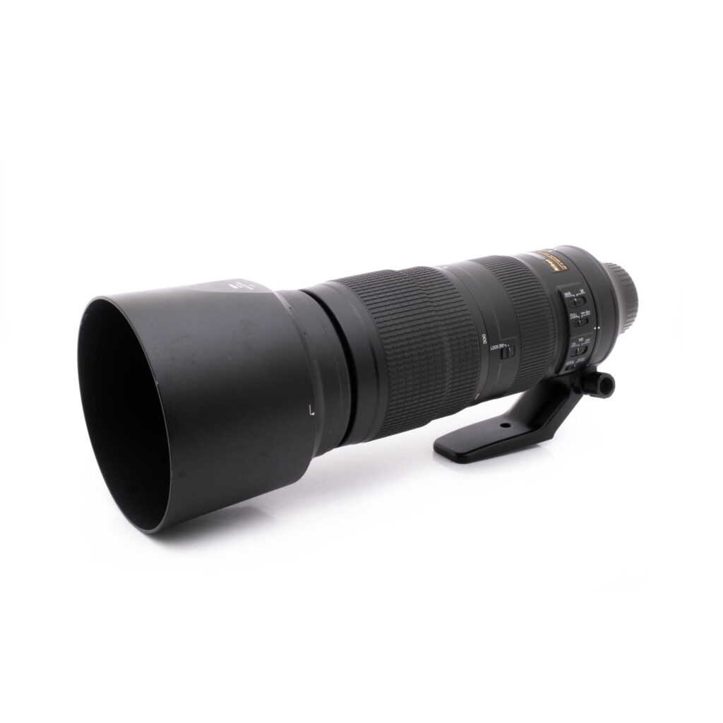 Nikkor 200-500mm-1 Nikon AF-S Nikkor 200-500mm f/5.6E ED VR - Käytetty teleobjektiivi, musta, vastavalosuoja ja jalustakiinnitys, näkyy valkoisella pohjalla hieman vasemmalle suunnattuna.