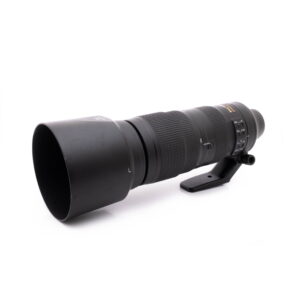 Nikkor 200-500mm-1 Nikon AF-S Nikkor 200-500mm f/5.6E ED VR - Käytetty teleobjektiivi, musta, vastavalosuoja ja jalustakiinnitys, näkyy valkoisella pohjalla hieman vasemmalle suunnattuna.