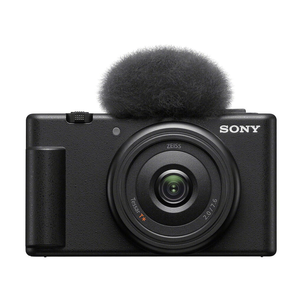 Sony_ZV-1F_008 Etukuva mustasta Sony ZV-1F -kompaktikamerasta, jossa on suuri pyöreä linssi ja siihen kiinnitetty sumea tuulilasi, joka on suunniteltu vähentämään melua äänitallennuksen aikana.
