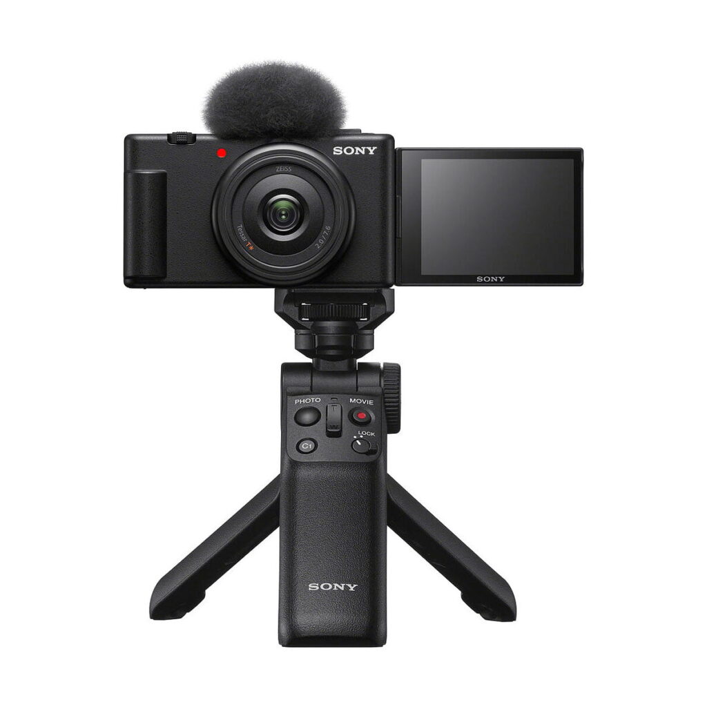 Sony_ZV-1F_009 Musta Sony ZV-1F -kompaktikamera, jossa on ulosvedettävä näyttö ja yläosan mikrofoni, asennettuna kompaktin Sonyn jalustakahvan varaan, valkoista taustaa vasten.