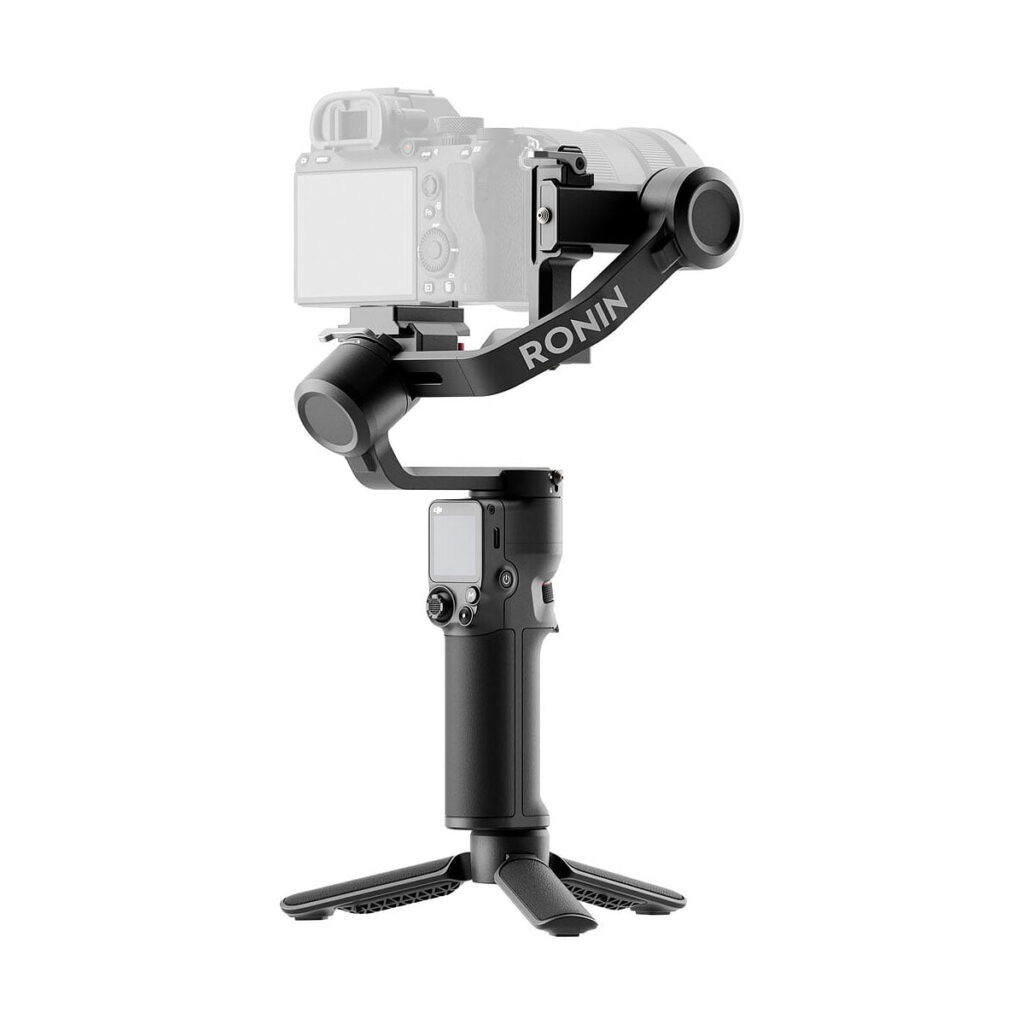 DJi_RS_3_Mini_002 The DJI RS 3 Mini gimbal, marked "RONIN", appears in black with a camera mounted at the top and a removable stand at the bottom. The camera appears slightly faded to highlight the gimbal stabilizer.