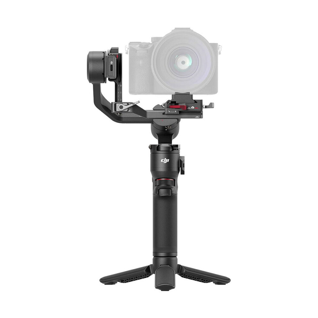 DJi_RS_3_Mini_003 The DJI RS 3 Mini gimbal, a handheld gimbal with a digital camera mounted on it, is shown on a small tripod against a white background. The handle features controls and a brand logo for ease of use.