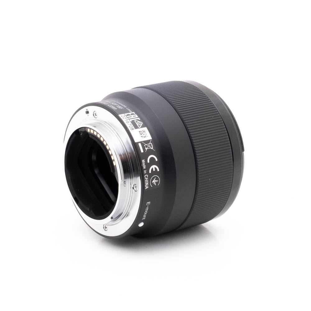 Sony 50mm f18-1 Lähikuva Sony FE 50mm f/1.8 - Käytetty -objektiivista kyljellään, jossa korostuvat metallinen kiinnitys elektronisilla koskettimilla, sertifiointimerkinnät ja piipun kuvioitu ote.