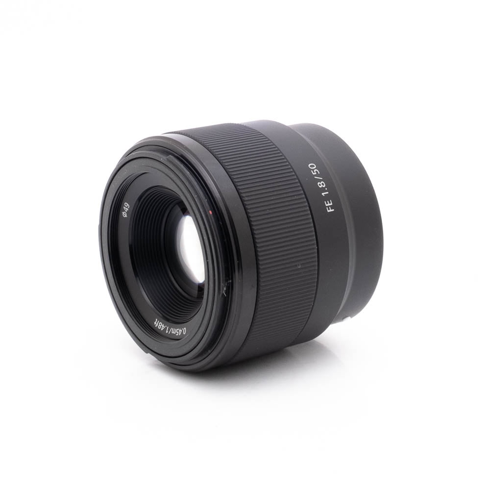 Sony 50mm f18-2 Sony FE 50mm f/1.8 - Käytetty -objektiivi, jonka kyljessä lukee "FE 1.8/50", on kuvattu valkoisella pohjalla, ja objektiivin etulasi ja objektiivin tynnyri on kuvakulmassa.
