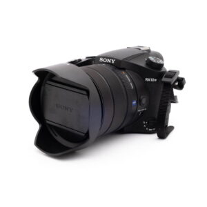 Sony RX10 IV-5 Musta Sony RX10 IV - Käytetty digikamera, jossa on suuri objektiivi ja vastavalosuoja, edestä vasemmalta valkoista taustaa vasten.