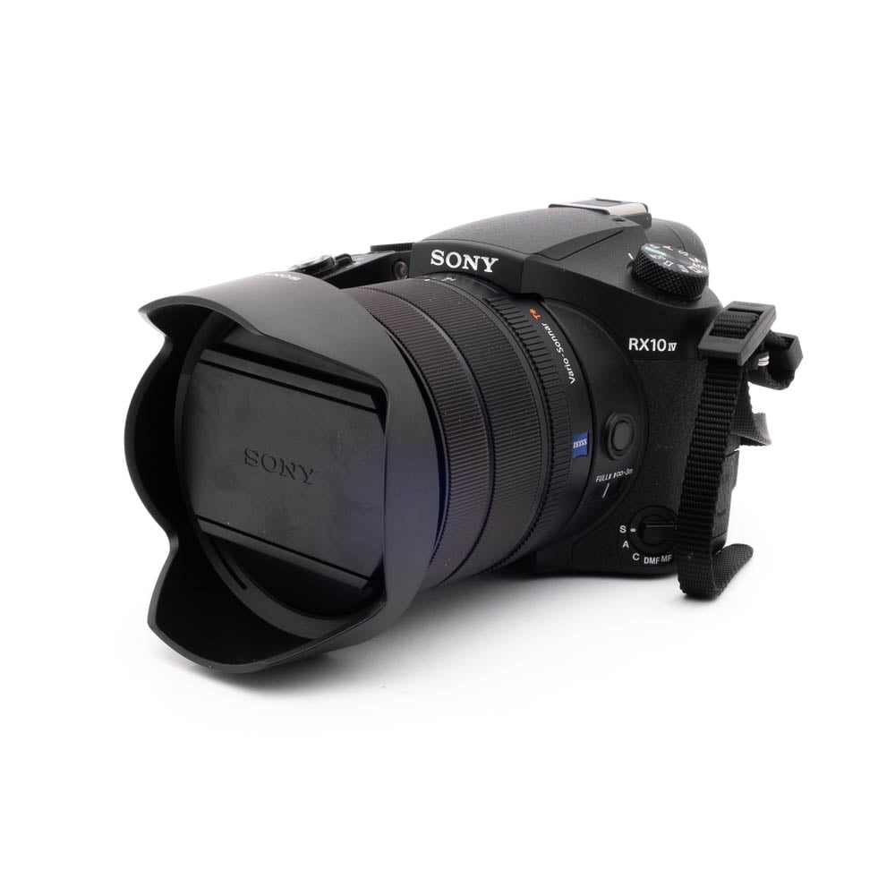 Sony RX10 IV-5 Musta Sony RX10 IV - Käytetty digikamera, jossa on suuri objektiivi ja vastavalosuoja, edestä vasemmalta valkoista taustaa vasten.