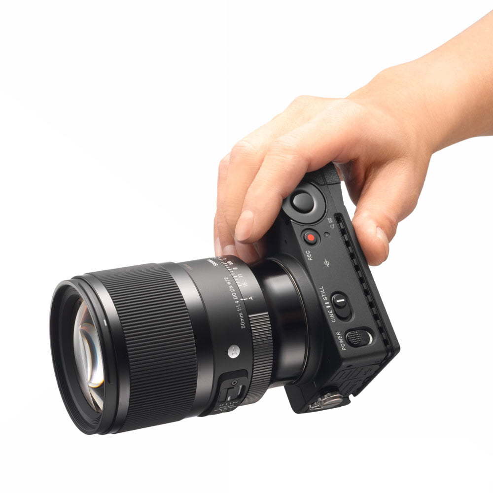 sigma_50mm_dg_dn_01 Kädessä on Sigma 50mm f/1.4 DG DN Art -objektiivi (Sony E) valkoista taustaa vasten - oikeutettu 150 euron käteispalautukseen.