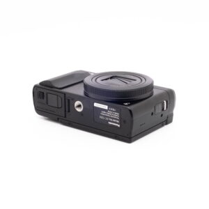 Panasonic DC-TZ90 7657-1 Panasonic Lumix DC-TZ90 - Käytetty kompakti musta digitaalikamera makaa selällään, jolloin sen alapuoli, jossa on tarrat, jalustakiinnike, akkulokero ja muistikorttipaikka, paljastuu valkoista taustaa vasten.