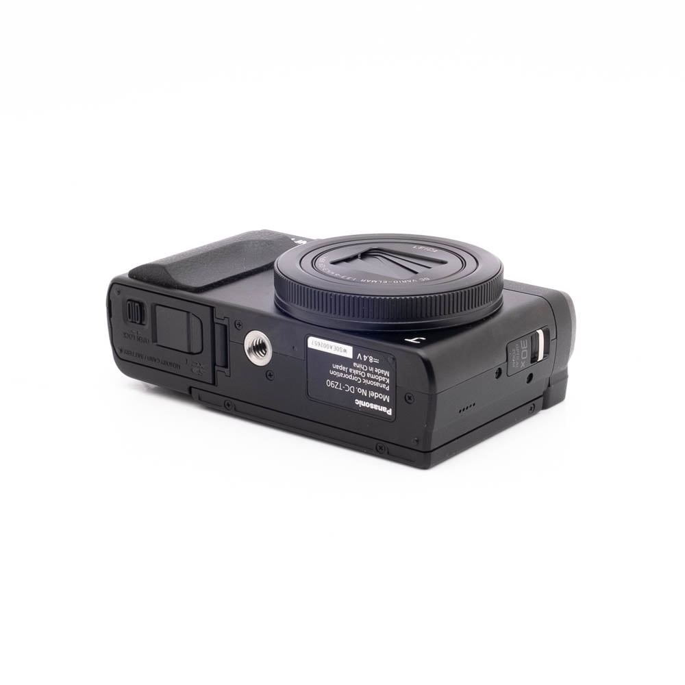 Panasonic DC-TZ90 7657-1 Panasonic Lumix DC-TZ90 - Käytetty kompakti musta digitaalikamera makaa selällään, jolloin sen alapuoli, jossa on tarrat, jalustakiinnike, akkulokero ja muistikorttipaikka, paljastuu valkoista taustaa vasten.
