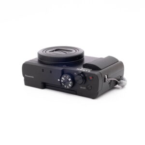 Panasonic DC-TZ90 7657-2 Musta Panasonic Lumix DC-TZ90 - Käytetty -digitaalikamera lepää valkoisella pinnalla, kun sitä katsotaan vinossa niin, että ylä- ja sivusäätimet, objektiivin suojus ja kiinnitetty rannehihna näkyvät.