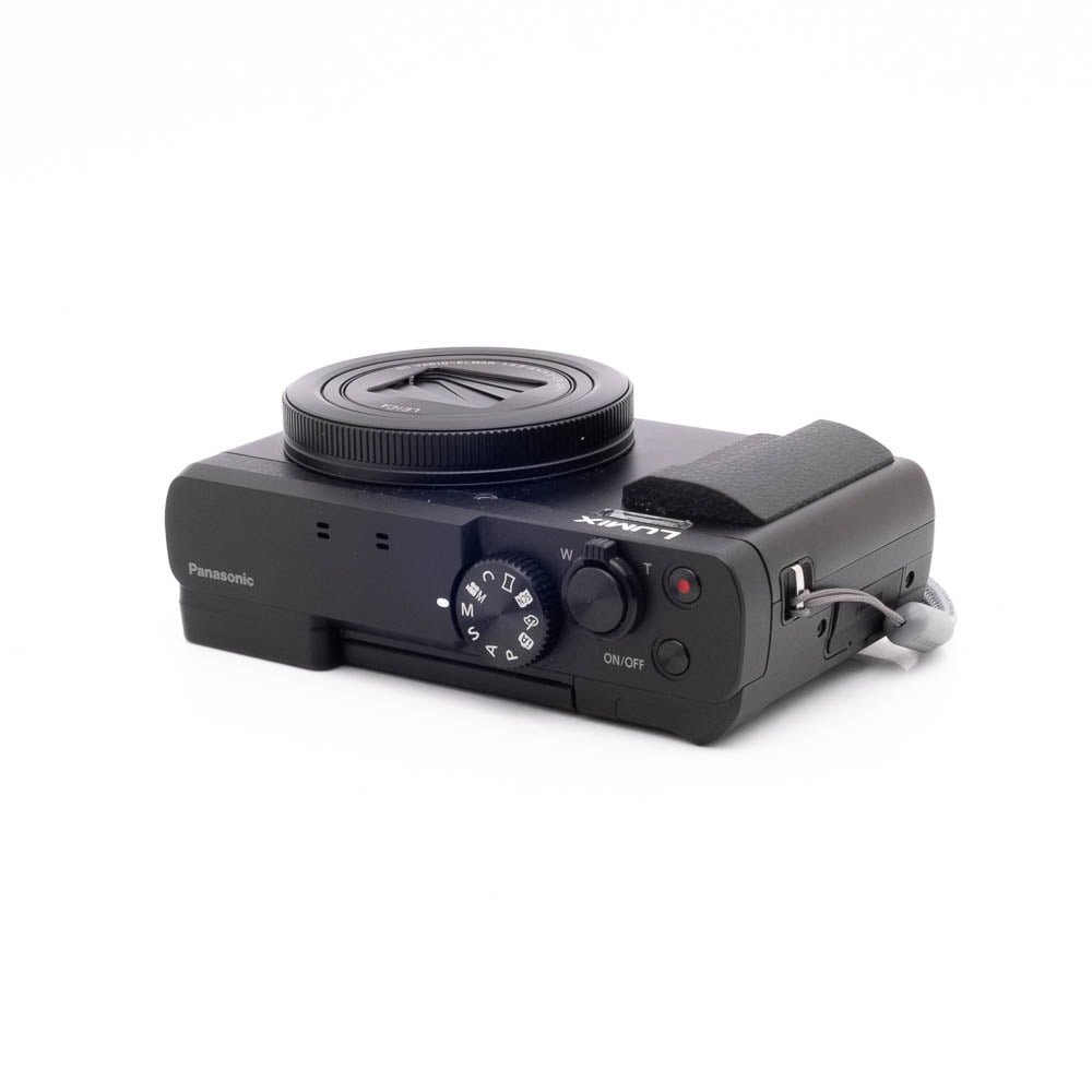 Panasonic DC-TZ90 7657-2 Musta Panasonic Lumix DC-TZ90 - Käytetty -digitaalikamera lepää valkoisella pinnalla, kun sitä katsotaan vinossa niin, että ylä- ja sivusäätimet, objektiivin suojus ja kiinnitetty rannehihna näkyvät.