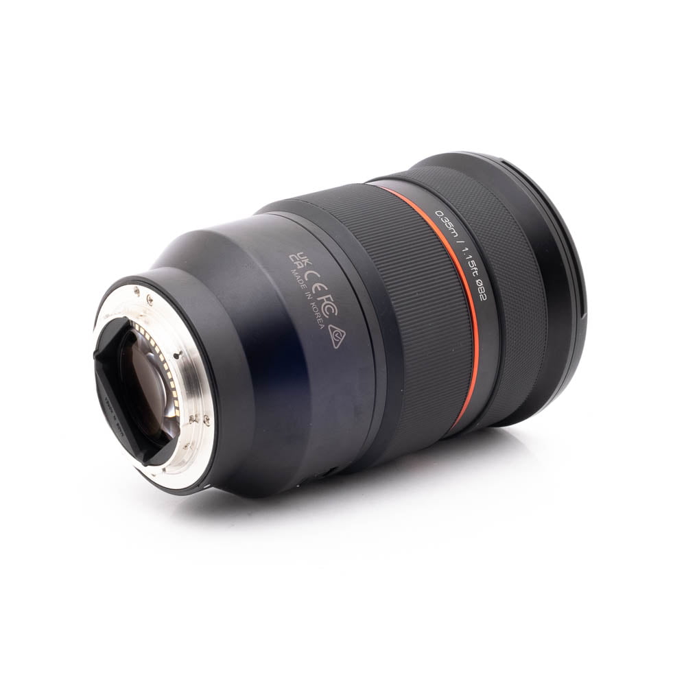 Samyang 24-70mm f28 FE Canon-1 Panasonic Lumix DC-TZ90 - Käytetty musta kameraobjektiivi, jossa on hopeinen metallijalusta ja punainen korostusrengas, kyljellään valkoista taustaa vasten.