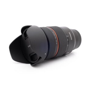 Samyang 24-70mm f28 FE Canon-3 Musta Panasonic Lumix DC-TZ90 - Käytetty kameran objektiivi, jossa on vastavalosuoja, punainen korostusrengas ja tuoteseloste, makaa kyljellään valkoista taustaa vasten.