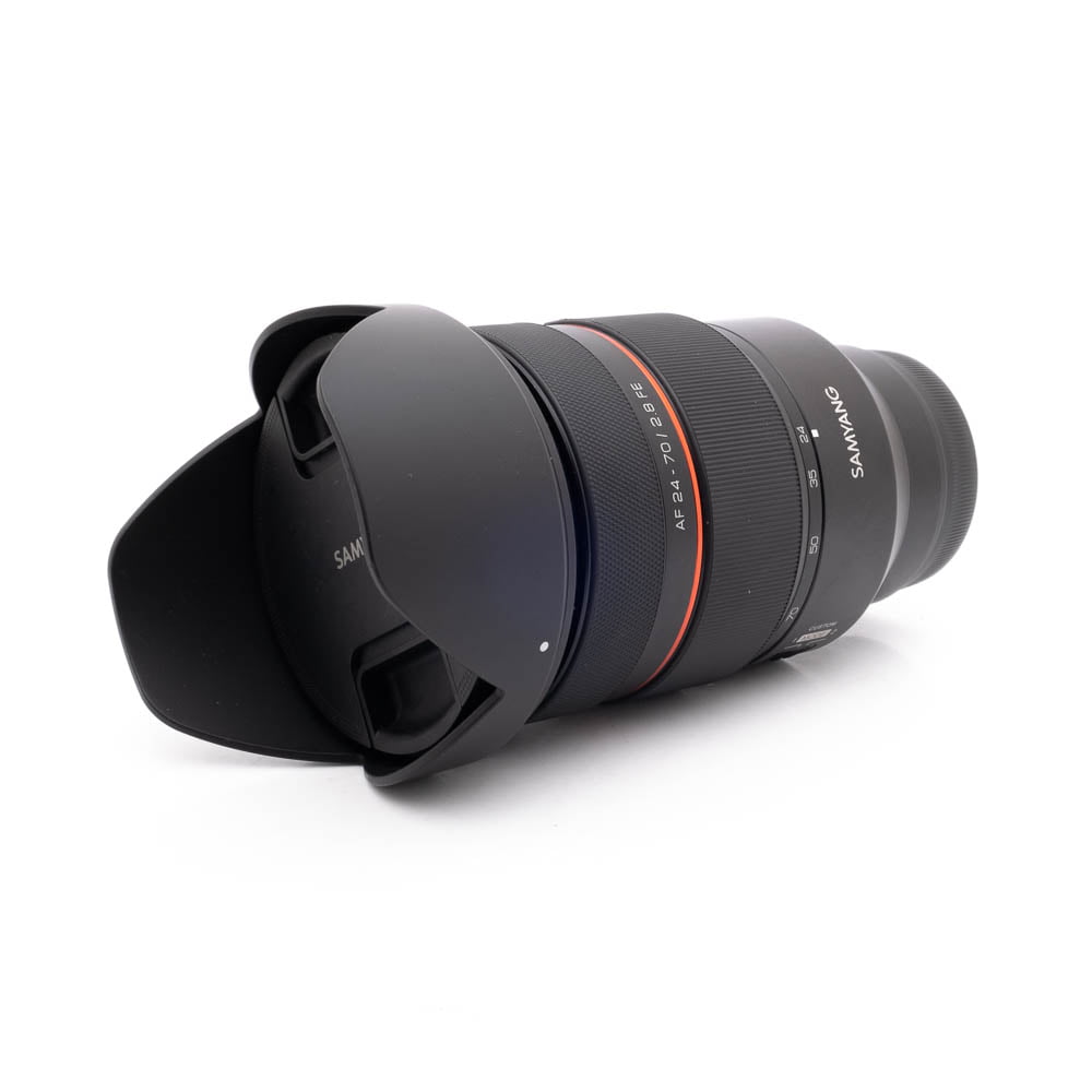 Samyang 24-70mm f28 FE Canon-3 Musta Panasonic Lumix DC-TZ90 - Käytetty kameran objektiivi, jossa on vastavalosuoja, punainen korostusrengas ja tuoteseloste, makaa kyljellään valkoista taustaa vasten.