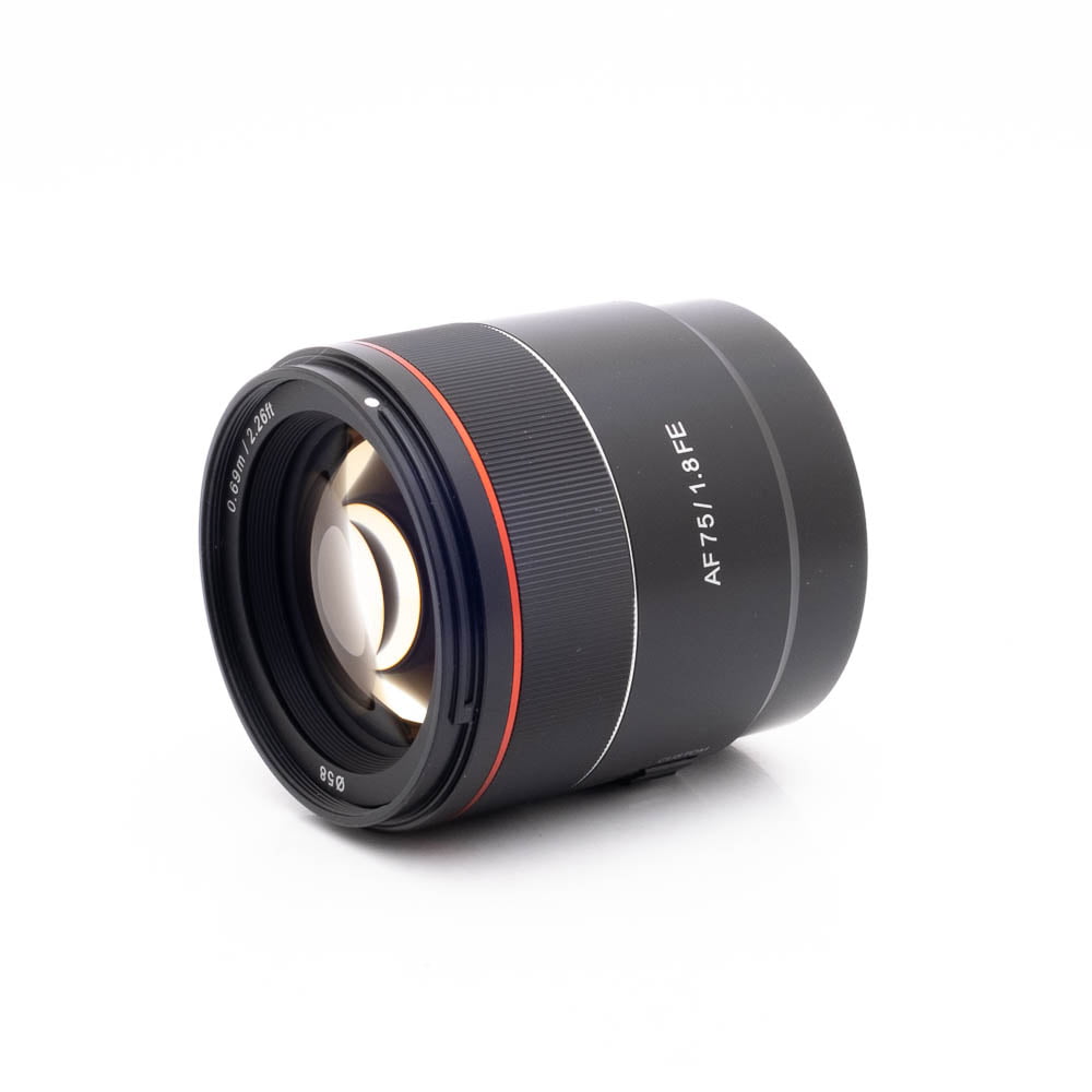 Samyang 75mm f18 FE Canon-2 Panasonic Lumix DC-TZ90 - Käytetty kamera on sijoitettu valkoiselle taustalle, ja sen objektiivin lasielementit näkyvät edessä.