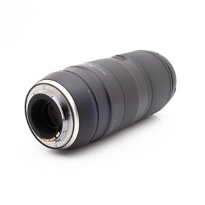 Tamron 100-400mm f45-63 Di VC USD Canon-1 Panasonic Lumix DC-TZ90 - Käytetty musta kameran zoom-objektiivi makaa kyljellään valkoista taustaa vasten, ja sen toisessa päässä on metallinen kiinnitys ja elektroniset kontaktit.