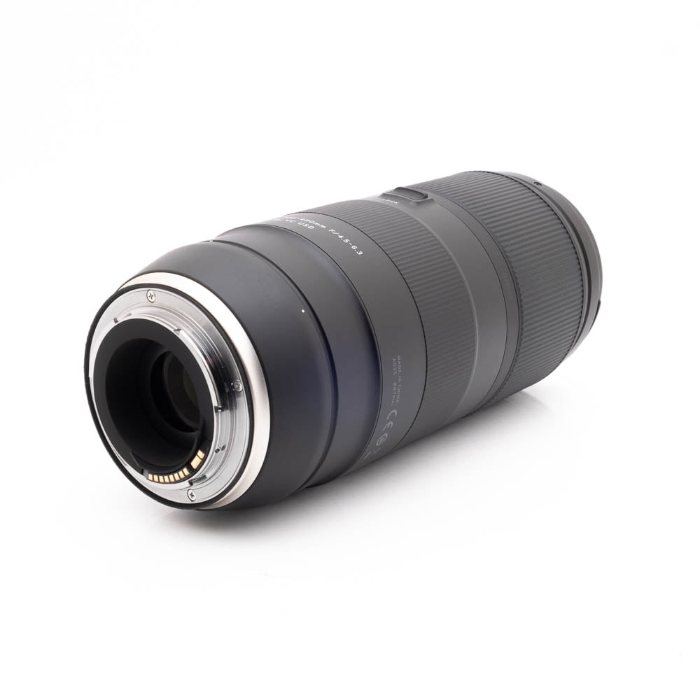 Tamron 100-400mm f45-63 Di VC USD Canon-1 Panasonic Lumix DC-TZ90 - Käytetty musta kameran zoom-objektiivi makaa kyljellään valkoista taustaa vasten, ja sen toisessa päässä on metallinen kiinnitys ja elektroniset kontaktit.