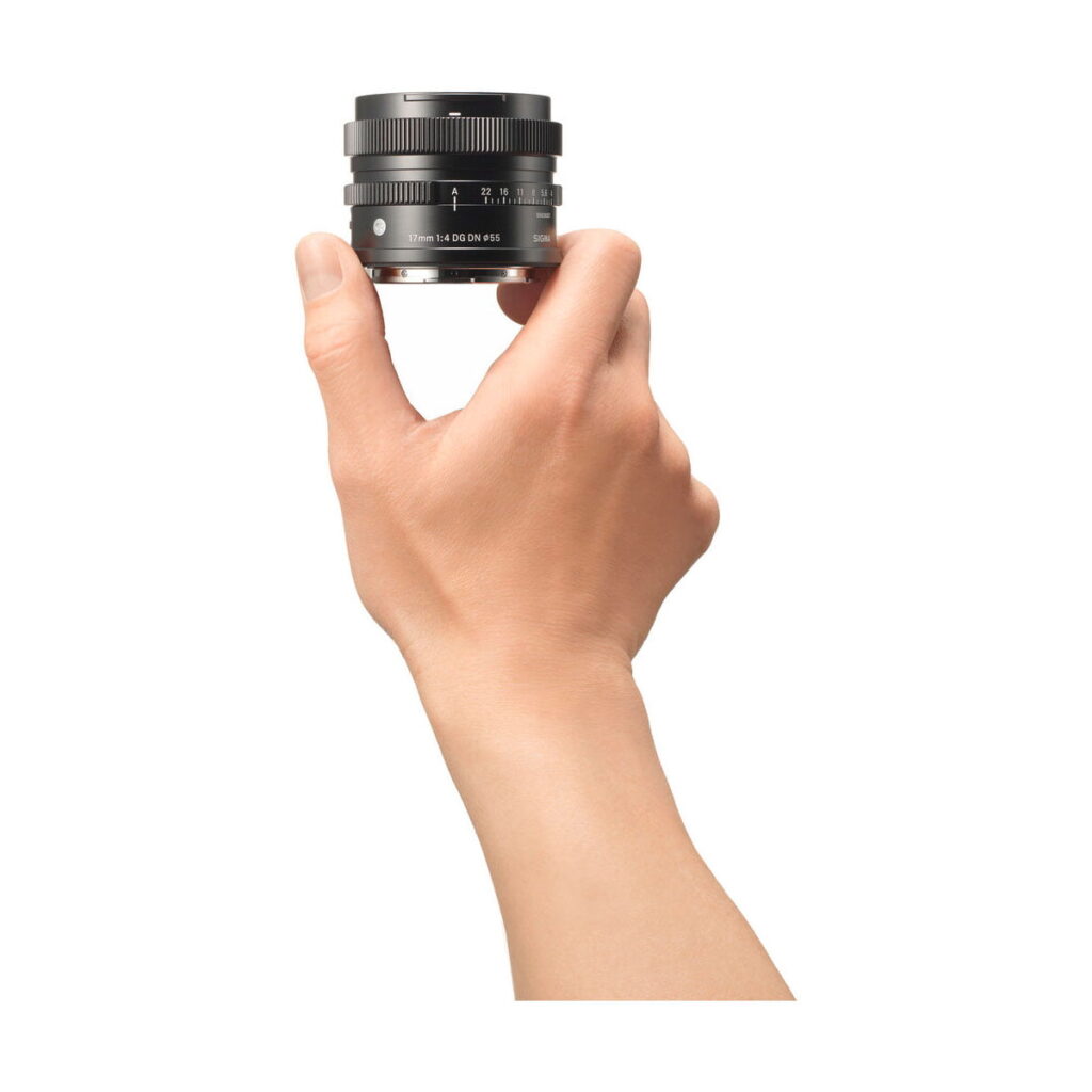 sigma_17mm_dg_dn Käsi pitää Sigma 17mm f/4 DG DN DN Contemporary -objektiivia Sonylle pystyssä valkoista taustaa vasten. Mustassa objektiivissa on näkyvät merkinnät ja säätörenkaat, ja käsi näkyy sivulta.