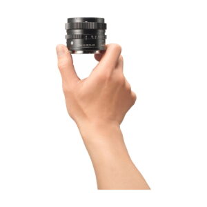 sigma_17mm_dg_dn Käsi pitää Sigma 17mm f/4 DG DN DN Contemporary -objektiivia Sonylle pystyssä valkoista taustaa vasten. Mustassa objektiivissa on näkyvät merkinnät ja säätörenkaat, ja käsi näkyy sivulta.