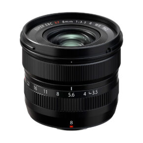 Fujifilm_XF_8mm_f_3_5_R_WR_001 Musta Fujifilm Fujinon XF 8mm f/3.5 R WR -objektiivi, jossa on kuvioidut tarkennusrenkaat ja aukkorenkaat, aukkoasteikko ja leveä etuelementti, on kuvattu valkoista taustaa vasten.