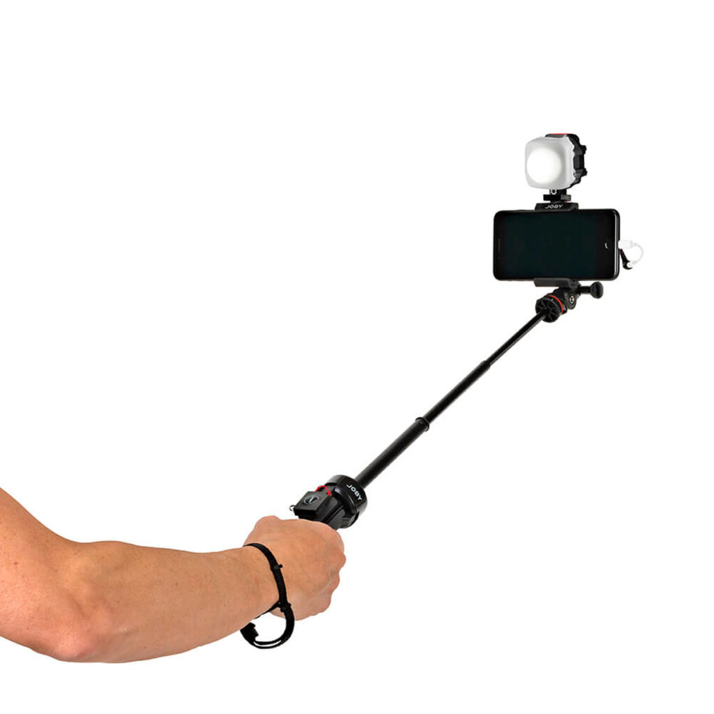 Henkilö pitää kädessään selfiekeppiä, johon on kiinnitetty älypuhelin, kiinnitetty Joby Beamo Mini LED-valo ja rannehihna. Keppi on ojennettu ja puhelin on henkilöstä poispäin.