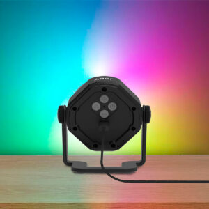 Joby-Beamo-Studio-Background-LED-Light-6 Joby Beamo Studio Background LED-valo istuu puupinnalla ja heijastaa sen takana olevalle seinälle sinisen, vihreän, vaaleanpunaisen ja keltaisen värisävyjä. Valosta lähtee virtajohto.