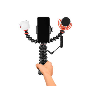 Joby-GorillaPod-Advanced-Mobile-Vlogging-Kit-2 Käsi pitää Joby GorillaPod Advanced Mobile Vlogging Kit - jossa on asennettu älypuhelin, LED-valo vasemmalla ja ulkoinen mikrofoni punaisella iskukiinnikkeellä oikealla - valkoisella taustalla.
