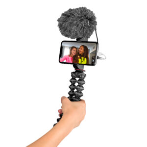 Joby-GorillaPod-Creator-Kit-3 Käsi pitelee Joby GorillaPod Creator Kitiä, joka tukee älypuhelinta, jossa on ulkoinen mikrofoni, ja näyttää näytöllä kaksi naista hymyilemässä selfieä tai videota varten.