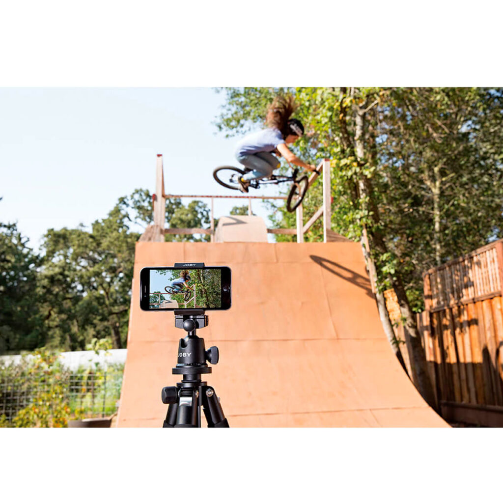 Joby-GripTight-Pro-Phone-Mount-6 Henkilö tekee BMX-temppua puisella rampilla ulkona, kun Joby GripTight Pro Phone Mount -puhelinpidikkeeseen kiinnitetty älypuhelin, joka on jalustalla, tallentaa toimintaa puiden ja puuaidan taustalla.