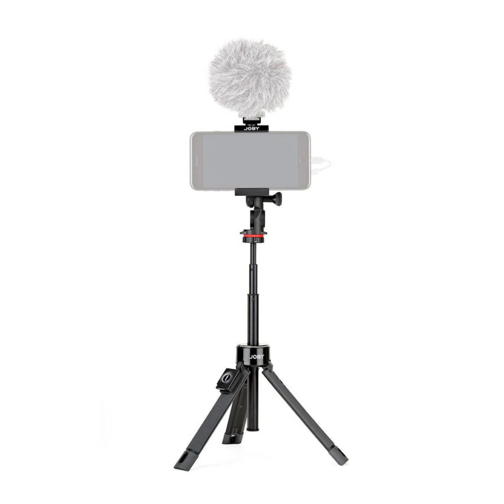 Joby-GripTight-Pro-TelePod-3 Älypuhelin on kiinnitetty täysin ulosvedettyyn mustaan Joby GripTight Pro TelePod -jalustaan, jossa on kolme jalkaa ja teleskooppinen keskipylväs, ja joka on varustettu ulkoisella mikrofonilla, jonka päällä on harmaa tuulisuoja.
