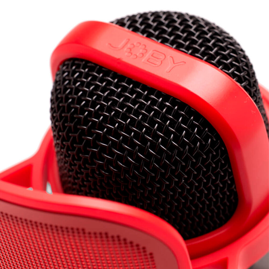 Joby-Microphone-Wavo-POD-4 Lähikuva Joby Wavo POD -mikrofoni, jossa on musta muotoilu ja metalliverkkosäleikkö, jota pidetään osittain kirkkaanpunaisessa pidikkeessä, jonka päällä on JOBY-brändinimi.