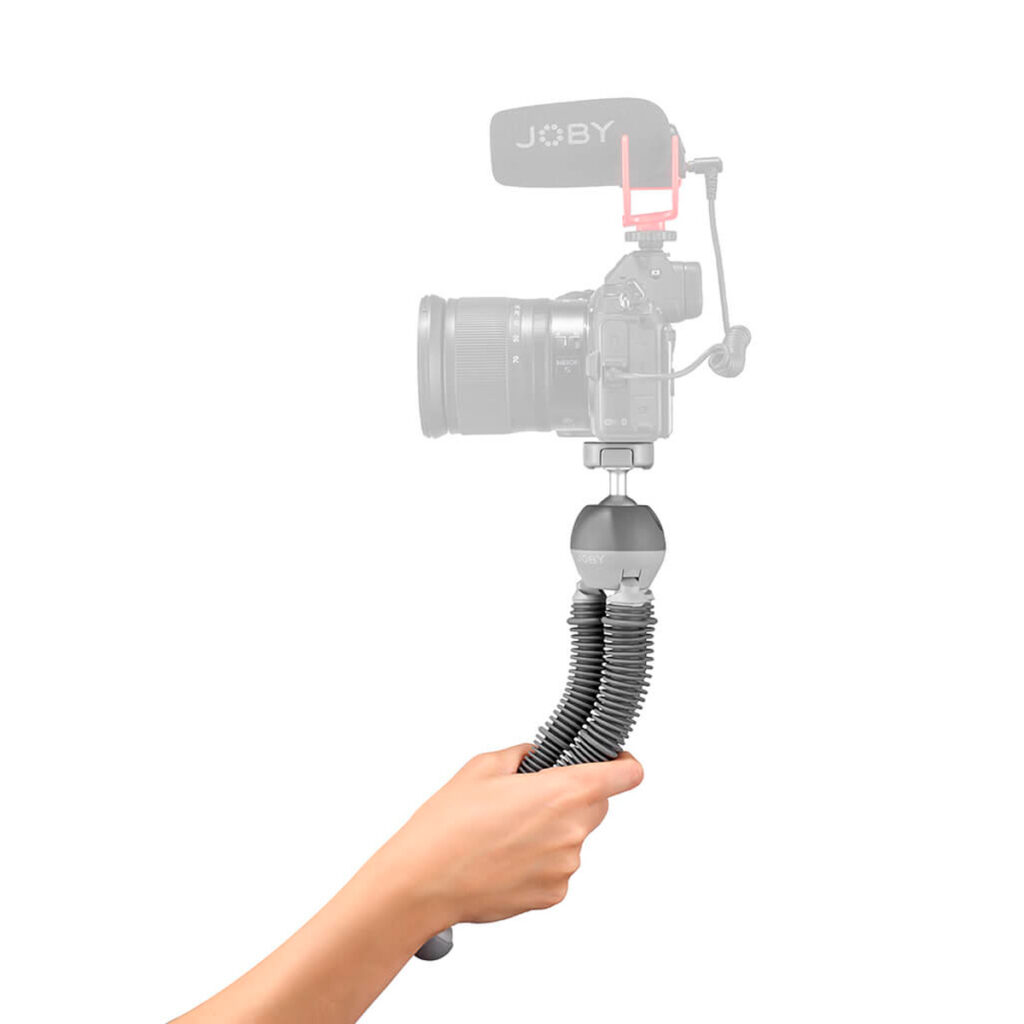 Kädessä on Joby PodZilla Flexible Tripod Large, jossa on segmentoidut mustat jalat ja kuulapääkiinnitys. Yläpuolella on häivytetty kuva kamerasta, johon on kiinnitetty mikrofoni, mikä havainnollistaa jalustan käyttötarkoitusta.