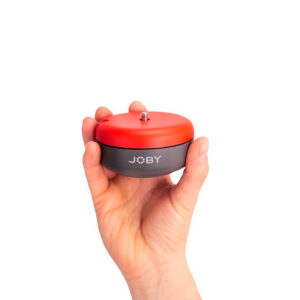 Joby-Spin-3 Kädessä on Joby Spin - Jalustapää, pieni, pyöreä laite, jossa on punainen yläosa, musta pohja ja ruuvikiinnitys kameran tai lisävarusteiden kiinnittämistä varten. JOBY-merkki näkyy valkoista taustaa vasten.