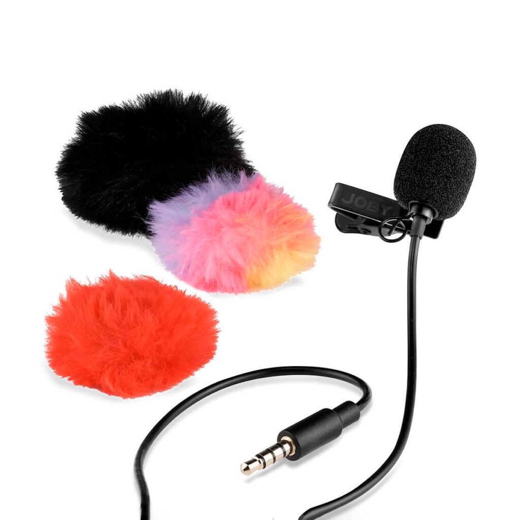 Joby-Wavo-Lav-Mikrofoni Joby Wavo Lav Mikrofoni, musta clip-on lavalier-mikrofoni, jossa on vaahtomuovinen tuulisuoja ja 3,5 mm:n liitin, on esillä kolmen karvaisen tuulisuojan vieressä: musta, punainen ja monivärinen sekoitus vaaleanpunaista, keltaista ja violettia.