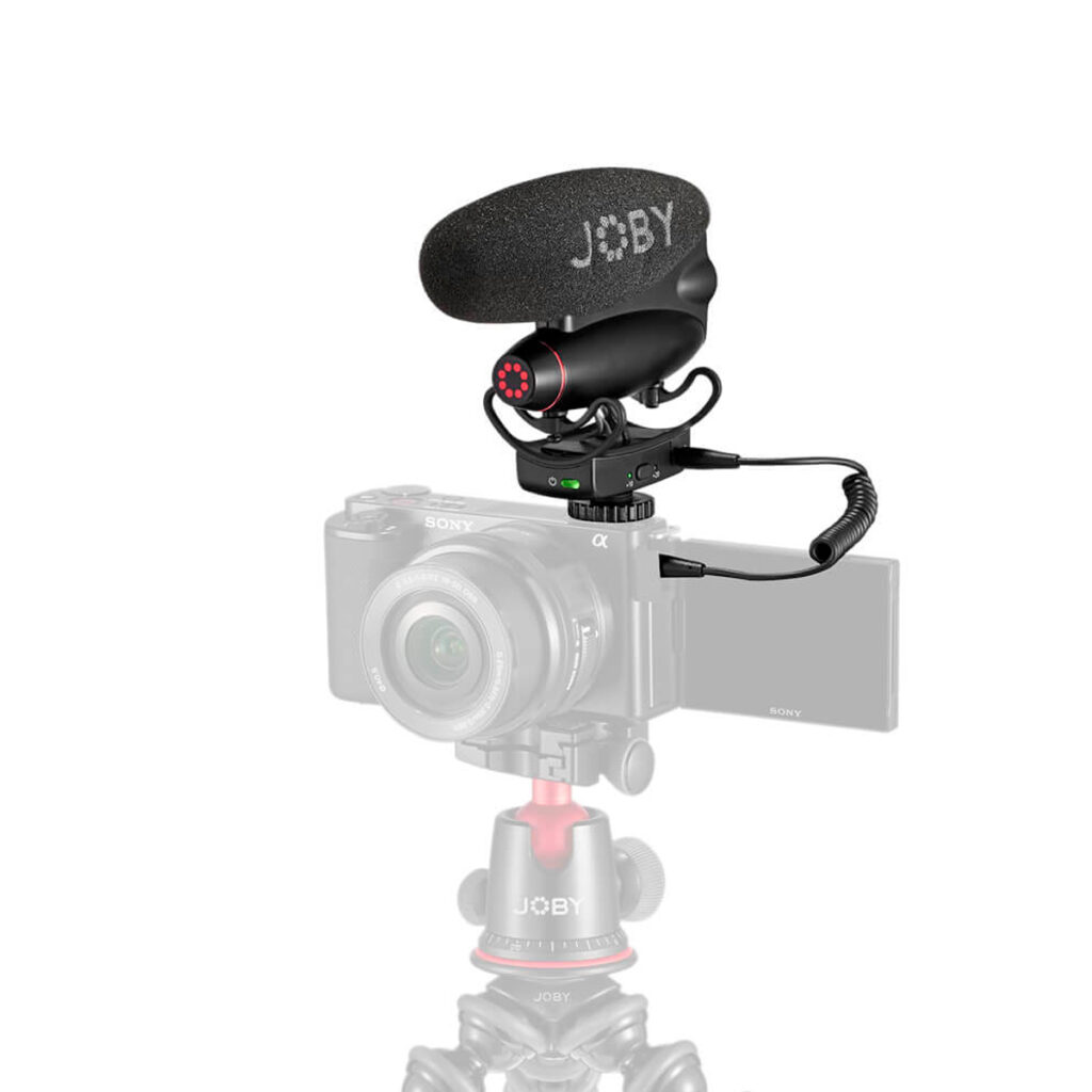 Joby-Wavo-Pro-DS-5 Pienikokoinen kamera, jossa on ulos käännettävä näyttö, on kiinnitetty Joby-jalustalle. Joby Wavo Pro DS - Mikrofoni on kiinnitetty kameran kuumakenkään kierrekaapelilla. Asetelma näkyy valkoista taustaa vasten.