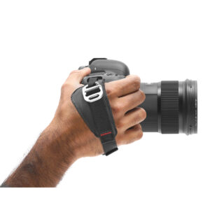 Peak-Design-Clutch-2 Henkilön käsi pitää mustaa DSLR-kameraa Peak Design Clutch -rannehihnan avulla, joka tarjoaa turvallisen otteen, kun kameran linssi on oikealle.