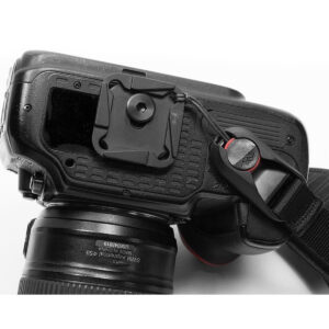 Peak-Design-Clutch-3 Lähikuva DSLR-kameran takaosasta ja sivusta, jossa on objektiivi, pikakiinnityslevy alla ja musta Peak Design Clutch -rannehihna kiinnitettynä ankkurilinkillä.