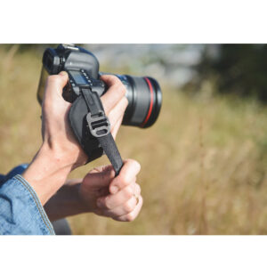 Peak-Design-Clutch-5 Farkkupaitainen henkilö pitelee DSLR-kameraa molemmilla käsillään ja säätää Peak Design Clutch -rannehihnan hihnaa ulkona auringonpaisteisella nurmikolla.