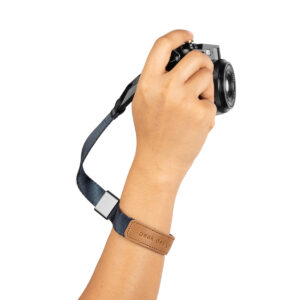 Peak-Design-Cuff-Midnight-2 Käsi pitää mustaa kameraa, jossa on Peak Design Cuff rannehihna, midnight-sininen rannehihna, jossa on ruskea nahkalaikka, eristettynä valkoisella taustalla.