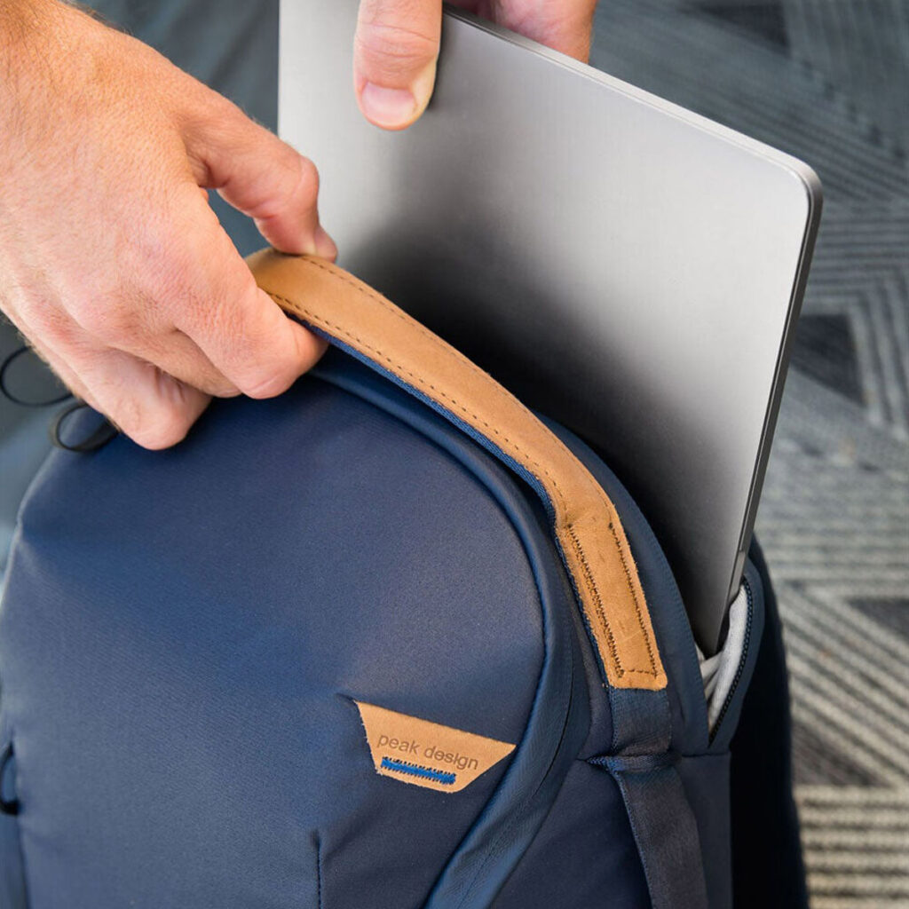 Peak-Design-Everyday-Backpack-Zip-20L-midnight-6 Henkilö laittaa hopeanvärisen kannettavan tietokoneen tummansinisen Peak Design Everyday Backpack Zip 20L -kamerareppu -repun pehmustettuun lokeroon, jossa on ruskehtava nahkakahva ja joka lepää kuviollisella matolla.