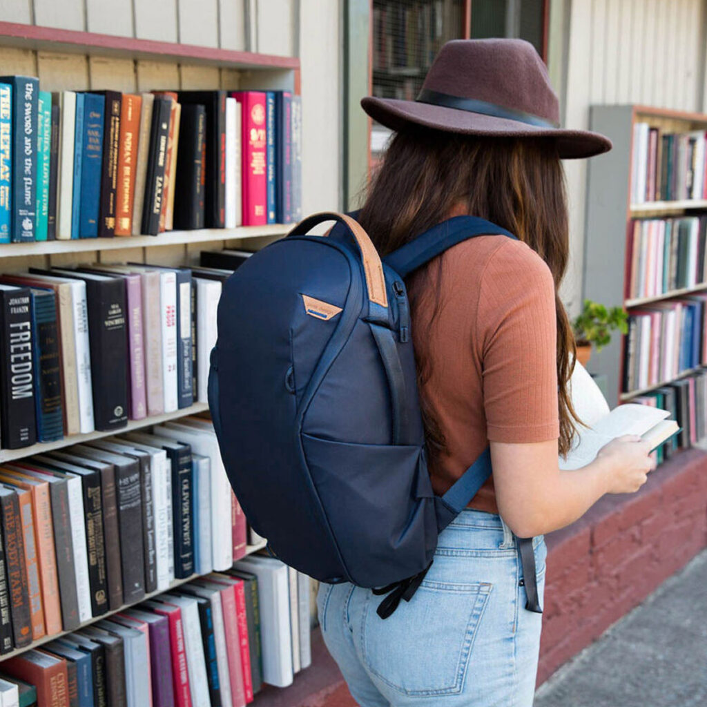 Peak-Design-Everyday-Backpack-Zip-20L-midnight-7 Ruskeaan hattuun, ruskeaan paitaan ja farkkuihin pukeutunut nainen, jolla on Peak Design Everyday Backpack Zip 20L -kamerareppu, tummansininen selailee ulkona olevaa kirjahyllyä, kädessään avoin kirja.