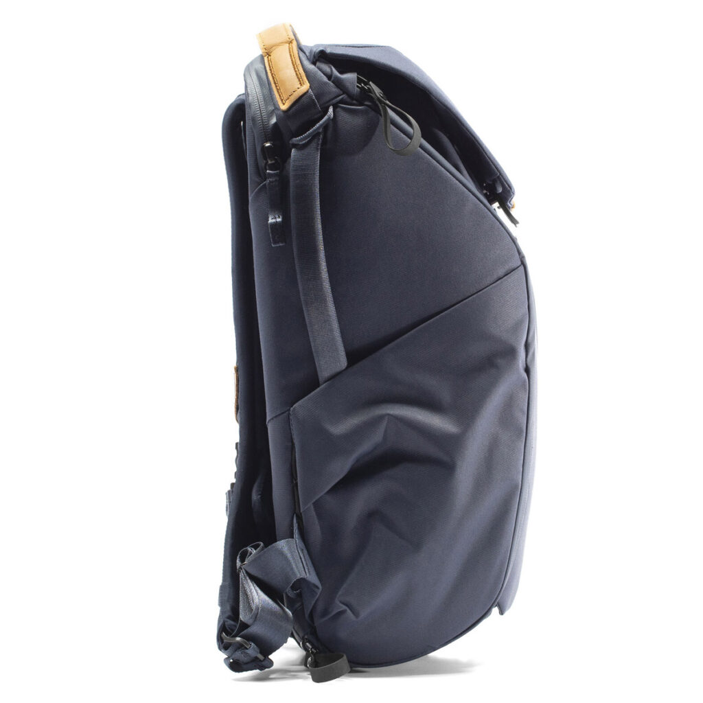 Peak-Design-Everyday-Backpack-v2-20l-midnight-4 Sivunäkymä Peak Design Everyday Backpack v2 20L -kamerareppu tummansinisessä värissä, jossa on ruskehtava nahkainen korostus yläkahvassa, näkyvät vetoketjut, pehmustetut olkahihnat ja tyylikäs, jäsennelty muotoilu hieman pyöristetyllä etuosalla.