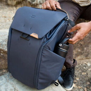 Peak-Design-Everyday-Backpack-v2-20l-midnight-6 Ulkona vaellusreitillä ruskeisiin housuihin ja mustiin kenkiin pukeutunut henkilö pitelee Peak Design Everyday Backpack v2 20L -kamerareppu, tummansininen -reppua toisella kädellä samalla kun hän laittaa mustan vesipullon sen sivutaskuun.
